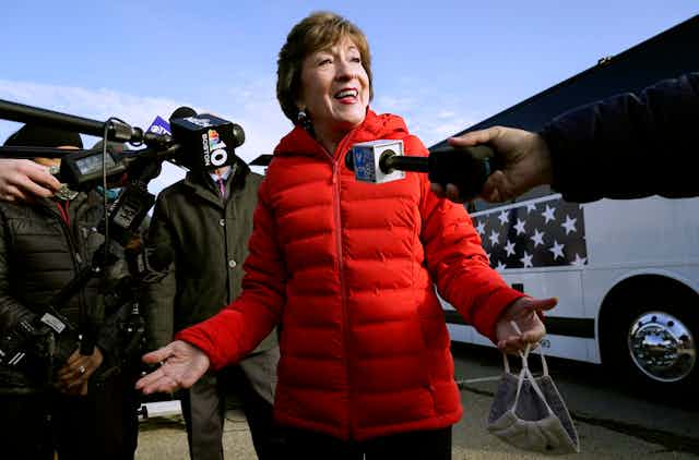 A smiling woman in a red puff jacket speaks into microphones.