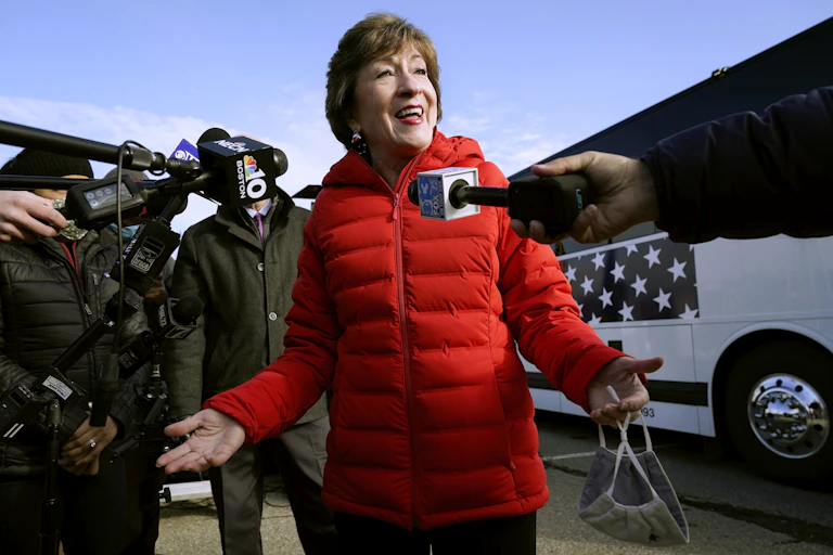 A smiling woman in a red puff jacket speaks into microphones.