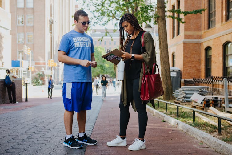 Two young people look toward a piece of paper, standing on what appears to be a college campus with open space and red brick buildings.