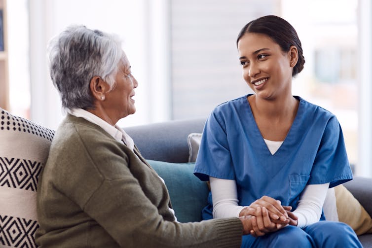 A young woman in scrubs sitting with an older woman, holding her hand.