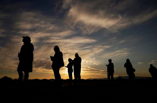 Silhouetted against a bright sky, people stand in line