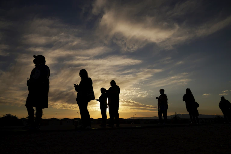 Silhouetted against a bright sky, people stand in line