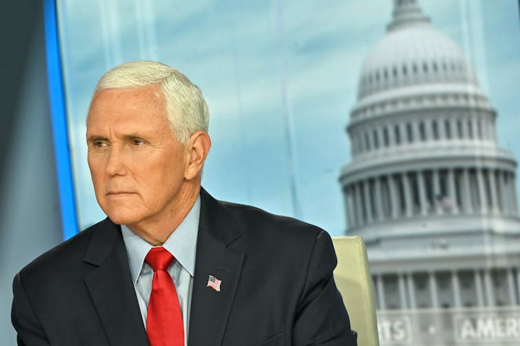 A middle-aged man with gray hair wearing a navy blue suit with red necktie sits in a television studio.