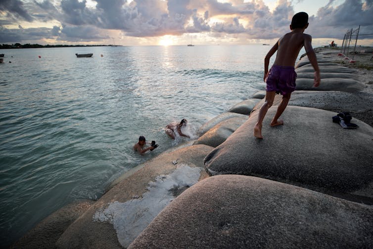 A child runs along large sandbags that form a sea wall while others pay in the water below.