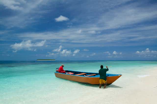 Two men in a fishing boat on a sandy beach in Tuvalu with islands in the background.