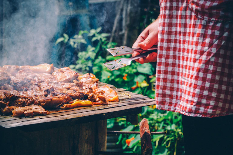 Person wearing a gingham apron holding tongs near cooked meat
