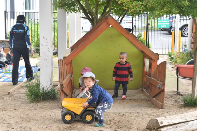 Children play outside in the sand, next to a log.