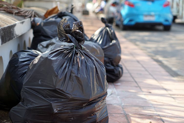 Uma pilha de sacos plásticos na calçada com um carro azul estacionado na estrada ao fundo.