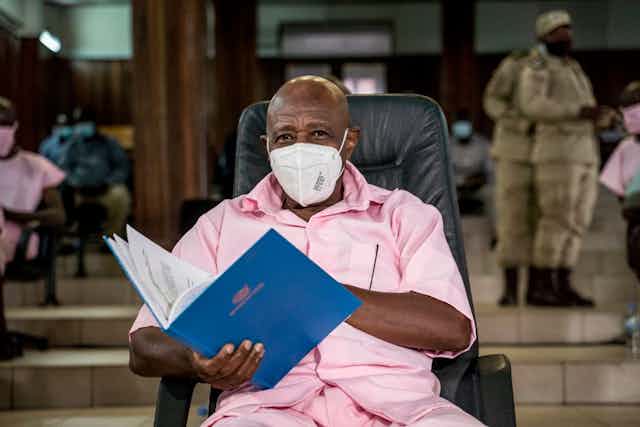 Man wearing a face mask and holding a case file while seated in court, dressed in a pink prison uniform.
