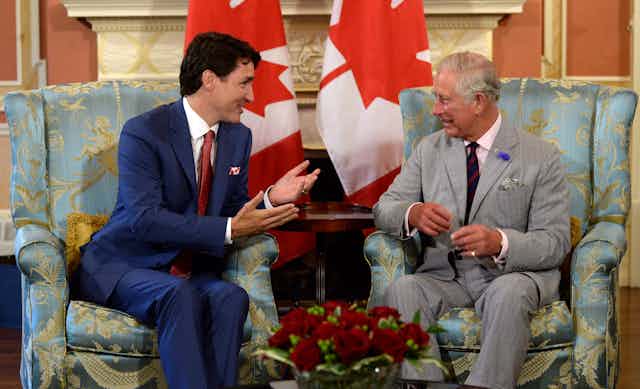 A dark-haired man in a blue suit sits in a chair talking to an older man with grey hair and a grey suit. Two Canadian flags are behind them.