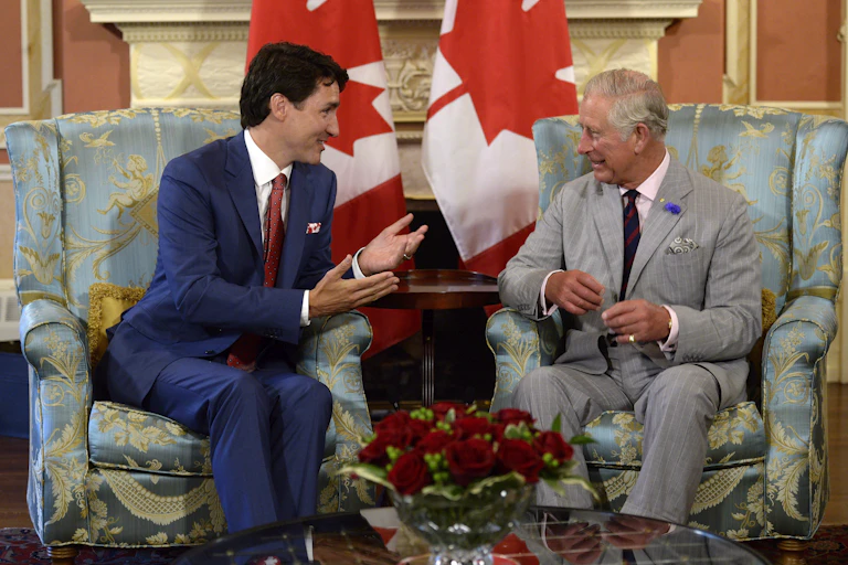A dark-haired man in a blue suit sits in a chair talking to an older man with grey hair and a grey suit. Two Canadian flags are behind them.