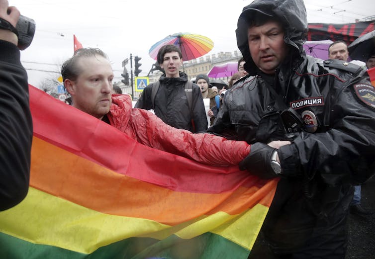 A man carrying a rainbow flag is pulled by the arm by a police officer.