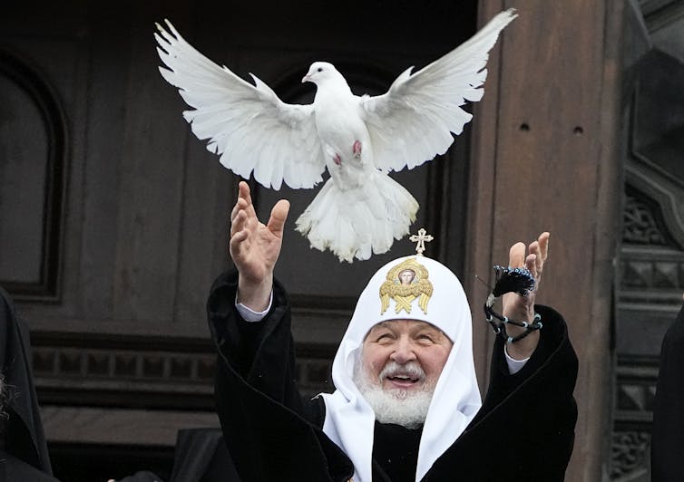 A man wearing white religious garb, including a white headpiece with a cross on it, releases a white dove with outstretched wings.