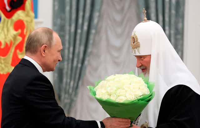 A balding man hands a bouquet or white roses to a man in a religious headpiece who smiles demurely.