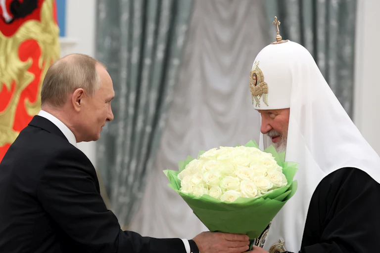 A balding man hands a bouquet or white roses to a man in a religious headpiece who smiles demurely.