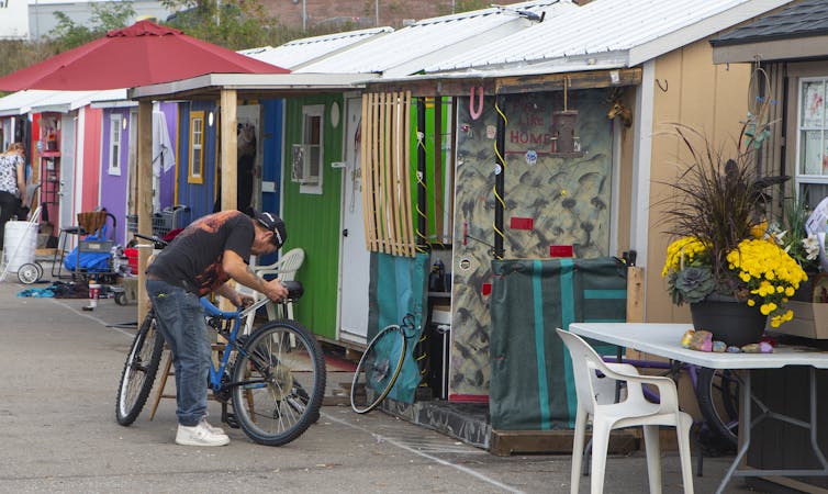 A man tends to his bicycle outside a row of small shelters.