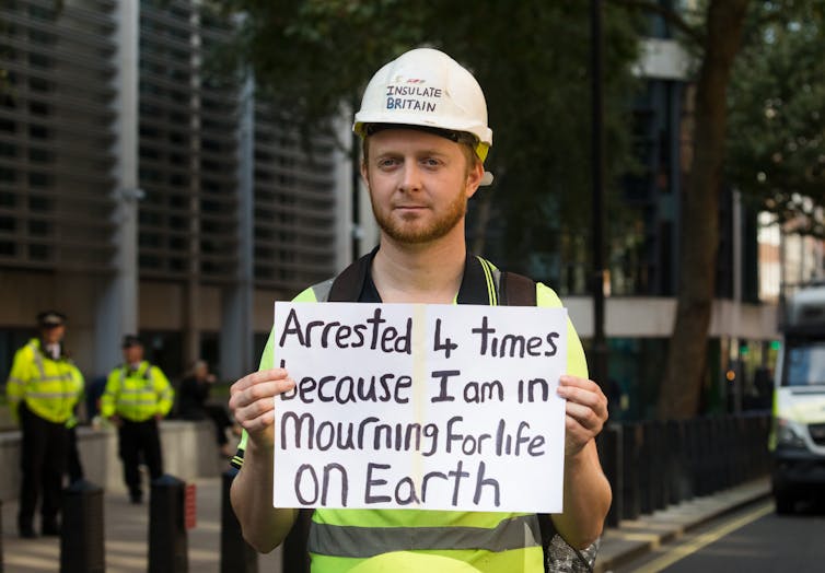 A man in a white construction helmet holds a placard reading 'Arrested 4 times because I am in mourning for life on Earth'