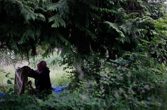 A man folds a blanket as he sits under the branches of a tree.