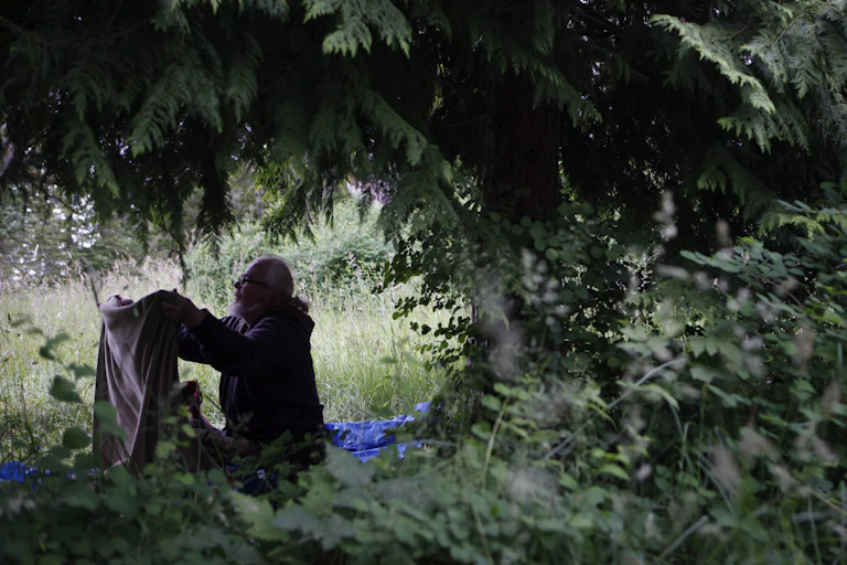 A man folds a blanket as he sits under the branches of a tree.