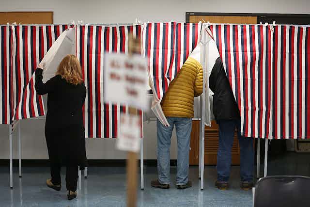Three people enter voting booths behind curtains with black, red, and white stripes.