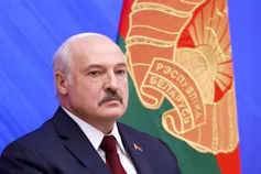 A bald man with a moustache is seen with an orange, red and green flag behind him.
