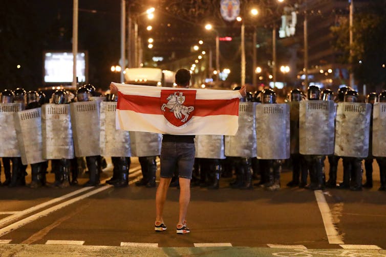 A man in shorts carries a white and red flag while standing in front of a line of police in riot gear.