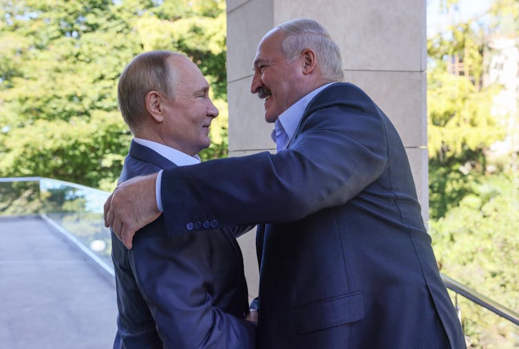 A balding man and another taller bald man with a moustache embrace.