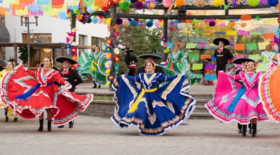Women in brightly colored dresses and men in large black hats dance in an outdoor plaza.