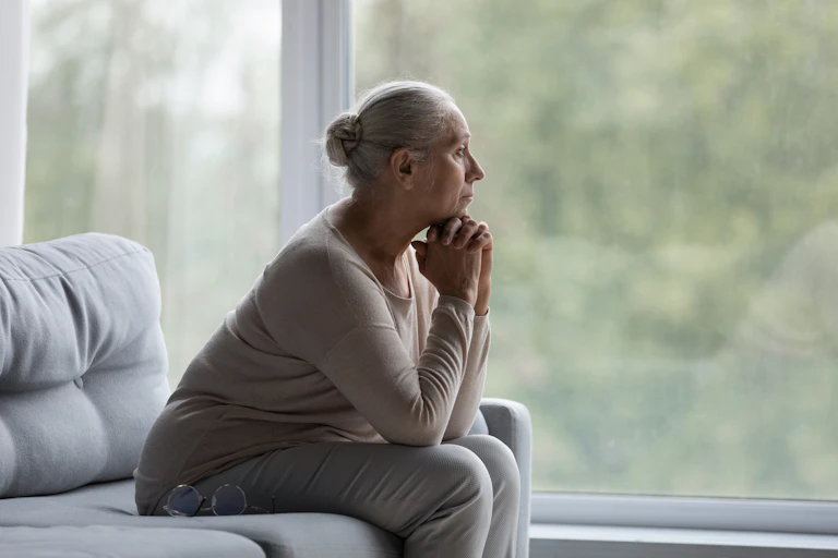 An older woman with her hair in a bun sitting on a sofa in front of a window with her chin on her hands