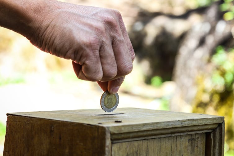 A hand placing a coin into a wooden box.