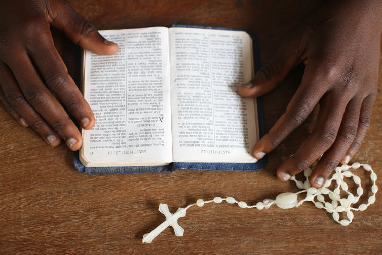 A photo taken above a table shows a person's hands holding an open Bible, with a rosary nearby.