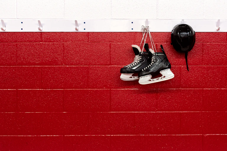 A pair of black hockey skates hang on a hook on a red and white wall.