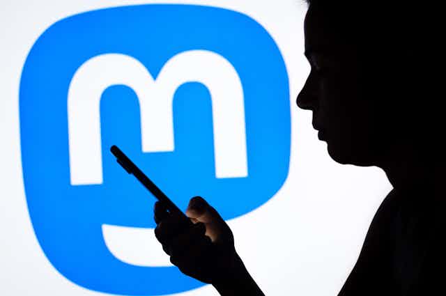 a photo illustration of the Mastodon logo seen in the background of a silhouette woman holding a mobile phone