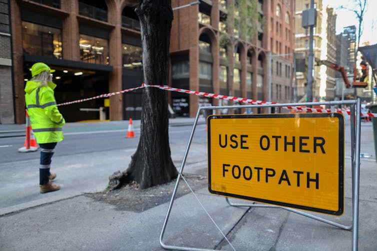 man in high-vis stands next to roped-off footpath with sign saying 'use opther footpath'