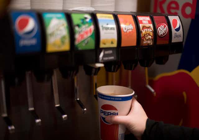 A soda fountain dispenser with eight choices, and a hand holding a paper Pepsi cup under one of the nozzles.