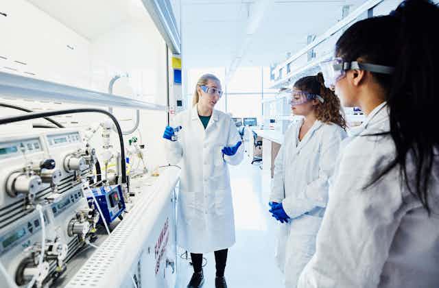 Three women in lab coats and goggles talk in a lab.