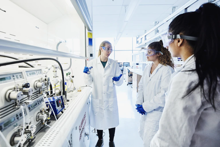 Three women in lab coats and goggles talk in a lab.