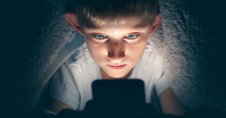 A young boy looking wide-eyed at a mobile phone under a blanket at night
