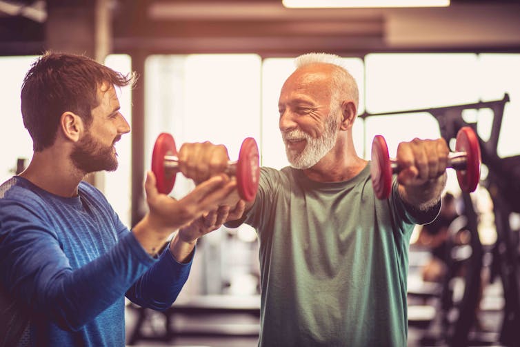 A young man assists a more senior man with weights at the gym.