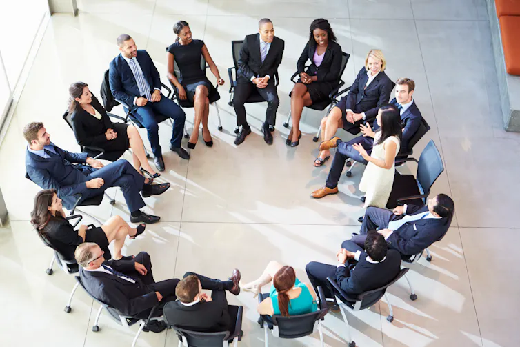 People sit in a circle while a woman stands addressing them.