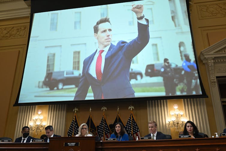 A man in a blue jacket, white shirt and red tie is shown raising his fist on a screen in a large meeting.