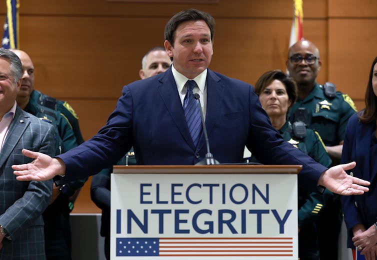 A white man dressed in a business suit stands with outstretched arms behind a lectern that has a sign bearing the words Election Integrity.