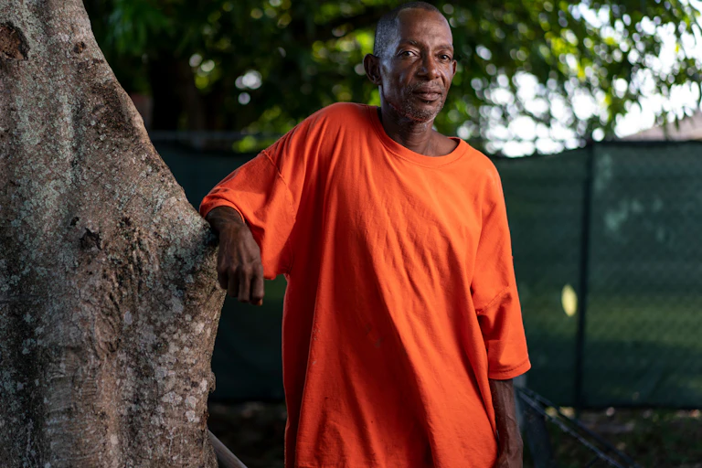 A black man is standing outside with his arm resting on a tree.