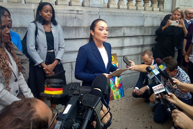 A woman in a blue pant suit and white shirt stands at a podium outside, facing a row of microphones at what appears to be a press conference.