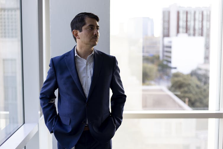 A Latino middle aged man wearing a blue suit stands in a modern looking room, with big glass windows, and looks to his left, with his hands in his pockets.