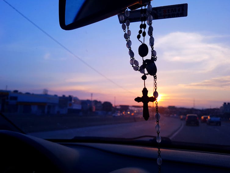 A rosary hangs from the rearview mirror of a car; background shows a sunrise