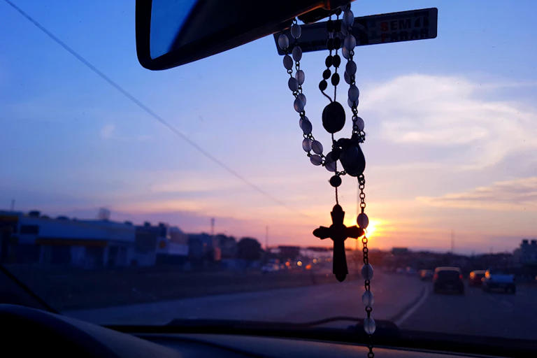 A scene looking out the front passenger seat of a car, with a rosary hanging from the rearview mirror, and a sunset ahead.