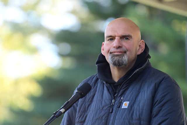 Wearing a dark blue jacket and standing before a microphone, U.S. Senate candidate John Fetterman speaks before supporters and the media at a political rally.