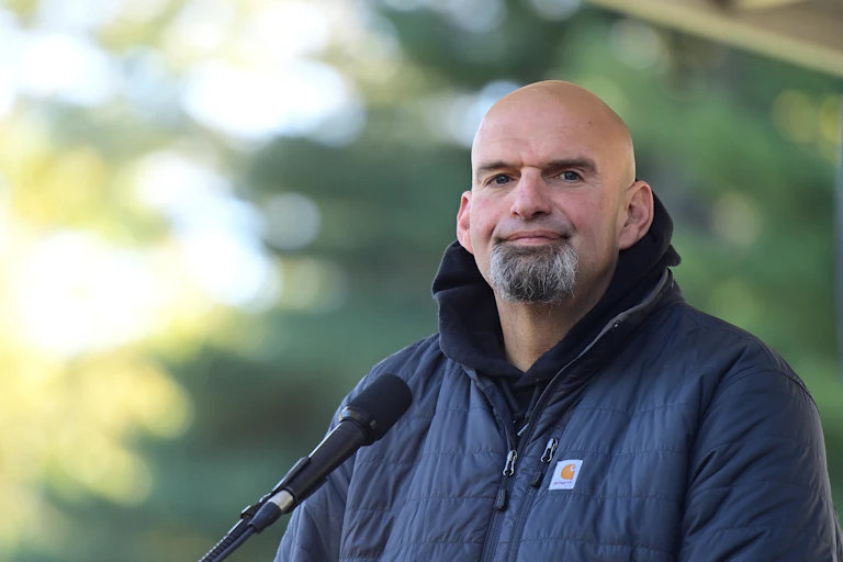 Wearing a dark blue jacket and standing before a microphone, U.S. Senate candidate John Fetterman speaks before supporters and the media at a political rally.