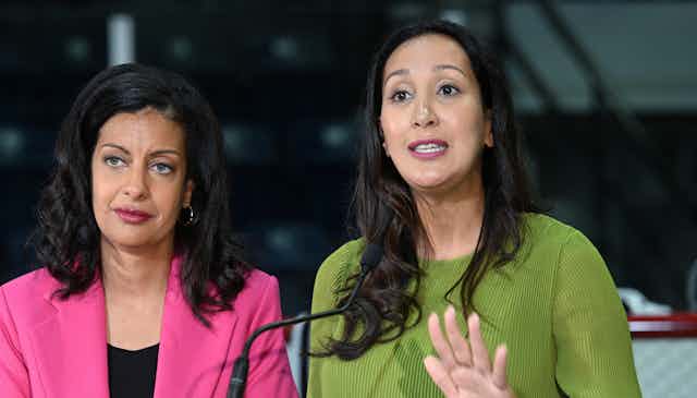 A woman with long dark hair wearing a green blouse speaks as a woman in a pink jacket with dark hair stands beside her.